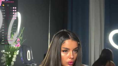 Media: A video of a young woman with long, straight, dark hair, styled with hairpins, wearing makeup, seated at a vanity table with a ring light, surrounded by floral arrangements and a computer screen displaying a video call.