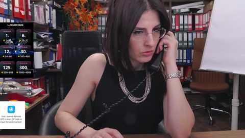 Media: Video of a pale-skinned woman with dark hair, wearing glasses and a black sleeveless top, sitting at a desk in a library.