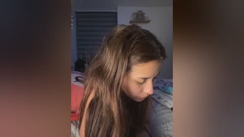 Media: Video of a young girl with long brown hair, wearing a red shirt, sitting on a bed with gray-patterned bedding in a dimly lit bedroom.