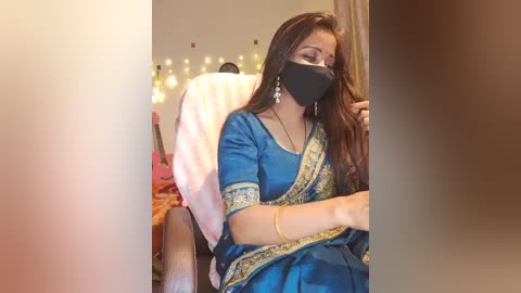 Media: A video shows a South Asian woman with long brown hair, wearing a blue sari with golden embroidery, black face mask, and silver earrings, seated in a cozy room with fairy lights and pink pillows.