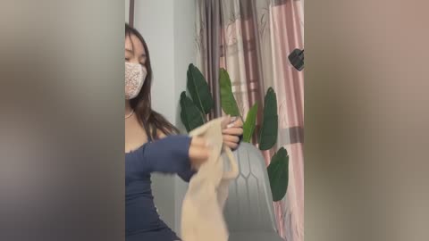 Media: A video of a woman with long brown hair wearing a face mask, a dark blue top, and a beige sweater, watering a potted plant with green leaves in a room with a pink curtain.