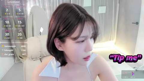 Media: A video of an Asian woman with short dark hair, wearing a white top, looking surprised in a modern bedroom with white curtains and a digital clock.