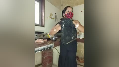 Media: A video shows a woman in a black dress, red face mask, and black headscarf preparing food in a rustic kitchen with a stove and sink.