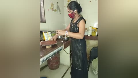 Media: Video of a woman in a traditional black dress and red mask, preparing food on a counter in a small, cluttered kitchen with white tiles and cooking utensils.