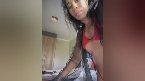 A video of a young Black woman with long black hair, wearing a red bra and black lace robe, standing in a bedroom with beige walls and wooden closet.