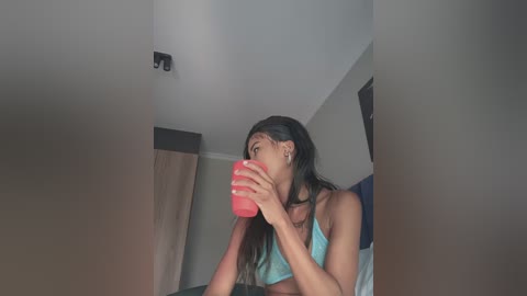 Video of a young woman with long black hair, wearing a blue tank top, drinking from a pink cup, indoors with muted colors and a minimalistic background.