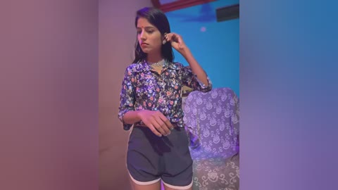 Media: Video of a young South Asian woman with medium skin tone, wearing a floral blouse and black shorts, adjusting her hair in a dimly lit room with blue and purple walls.