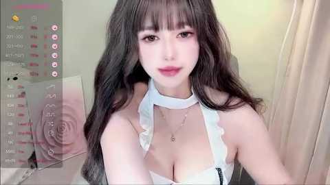 Media: Video of an East Asian woman with long, wavy black hair and fair skin, wearing a white halter top, smiling slightly. Background includes a calendar and floral decor.