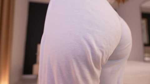 Media: A close-up video of a person's buttocks in white pajama pants, showing the fabric's texture and skin tone. The background is a blurred indoor setting with beige curtains and a black door.