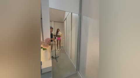 Media: Video of a modern, minimalist hallway with a tall mirror reflecting two women, one holding a camera, wearing casual clothes and standing in front of a white wall.