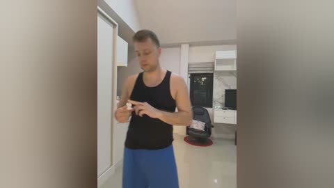 Media: Video of a fit, light-skinned man in a black tank top and blue shorts, adjusting his watch in a modern, minimalist kitchen with white cabinets, black office chair, and large windows.