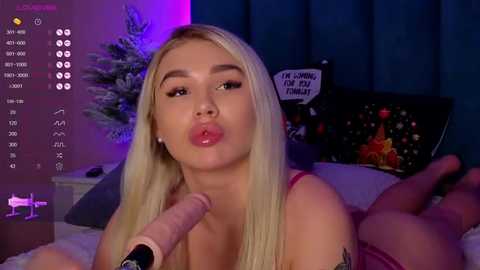 Media: Video of a blonde woman with fair skin, wearing a pink top, sucking a large, erect penis. She has a tattoo on her right arm. The room has a Christmas tree, blue curtains, and a black keyboard.