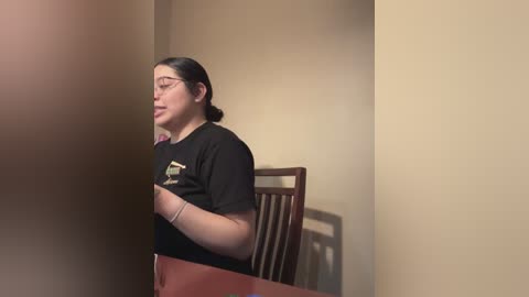 Media: Video of a young woman with medium brown skin, dark hair in a ponytail, wearing glasses and a black t-shirt, seated at a wooden table in a beige-walled room.