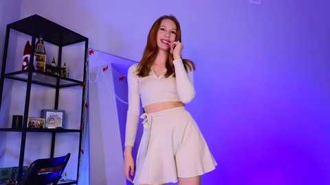 Media: Video of a smiling Asian woman with long brown hair, wearing a white crop top and high-waisted skirt, standing in a room with blue lighting, black shelves with framed pictures and collectibles, and a black chair.