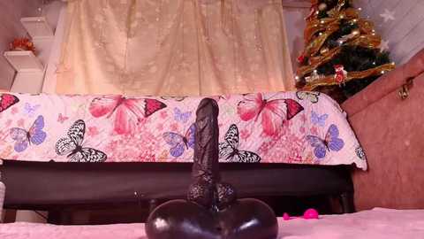 A video of a black dildo with a realistic cock and balls on a bedspread featuring pink butterflies and a Christmas tree in the background.