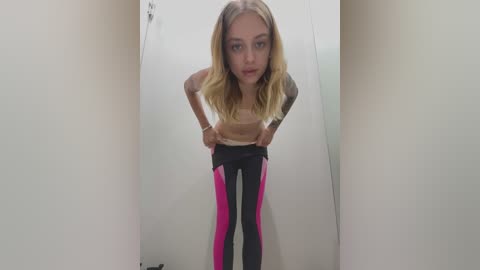 Media: Video of a slender, fair-skinned woman with shoulder-length blonde hair, wearing a beige tank top and black and pink leggings, bending over in a mirrored changing room.