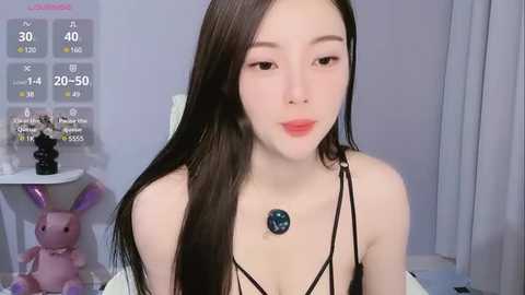 Media: Video of an East Asian woman with long black hair, pale skin, and light makeup, wearing a black strappy top, necklace, and sitting indoors. Background includes a weather forecast and a purple bunny statue.