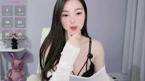 Media: A video of a young East Asian woman with long black hair, wearing a white shirt and black lingerie, seated indoors with a pink bunny toy, and a calendar in the background.