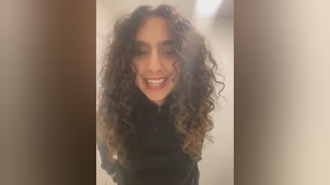 Media: Video of a smiling woman with curly, dark brown hair and light skin, wearing a black top, against a blurred, beige background.