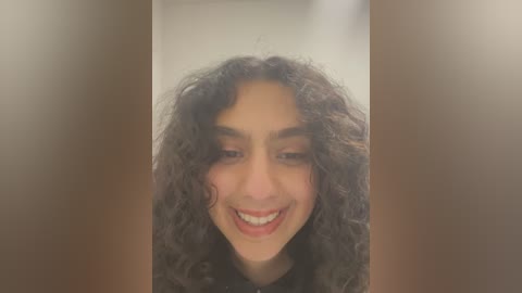 Media: Video of a young woman with curly, dark hair and light brown skin, smiling warmly, set against a blurry, neutral-toned background.