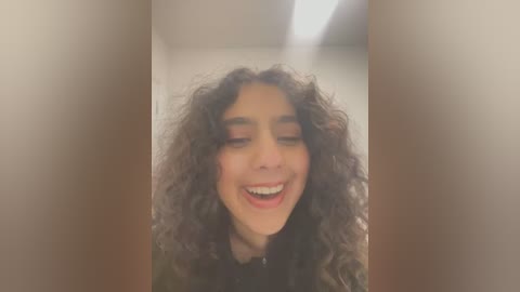 Media: A video of a young woman with curly, dark hair, light brown skin, and a joyful expression, captured from the waist up in a dimly lit hallway.