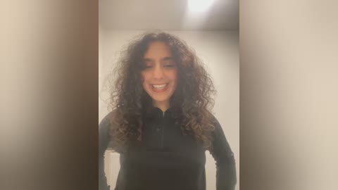 Media: Video of a smiling woman with curly, voluminous hair, wearing a black long-sleeved top, standing in a dimly lit room with soft, diffused lighting.