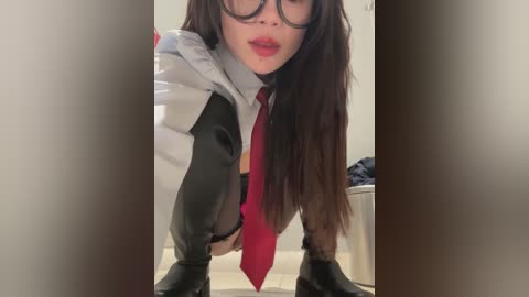 Media: Video of an Asian woman with long black hair, wearing glasses, a red tie, black leather boots, and a white and black jacket, squatting in a bathroom.