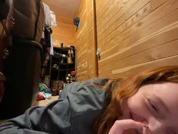 Media: Video of a young redhead girl with fair skin, wearing a blue shirt, lying on her side in a cozy, wooden-paneled room with a cluttered bookshelf in the background.