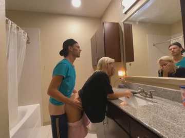 Media: Video of a muscular man in a blue shirt and blue shorts, standing behind a blonde woman in a black shirt, in a bathroom.