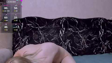 Video of a nude, fair-skinned woman lying on a black marble-patterned bedspread, partially visible in a dimly lit room with a digital overlay showing health statistics.