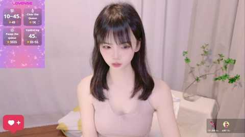 Media: Video of a fair-skinned East Asian woman with straight black hair and bangs, wearing a beige top, in a modern room with a potted plant.