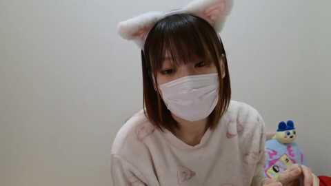 Media: Video of an Asian woman with shoulder-length dark hair wearing bunny ear headbands, a white mask, and a pink fuzzy sweater. She holds a plush toy in her right hand.