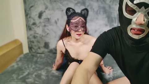 Media: Video of a light-skinned woman with long brown hair, wearing a black lace mask and revealing black lingerie, kneeling on a gray floor. A man in a black mask with silver eye holes, black shirt, and pants is behind her.