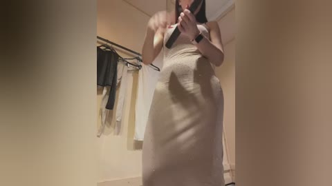 Video of a woman with light skin and long dark hair, wearing a tight, beige, strapless dress, holding a phone, in a beige-walled room with hanging clothes.