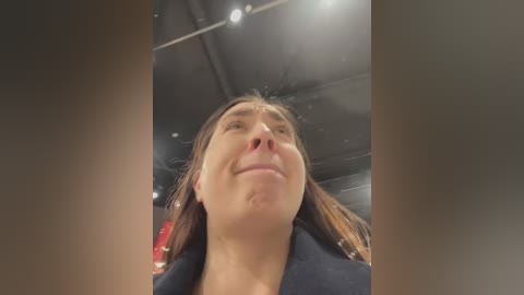 A video of a young woman with straight, shoulder-length brown hair, wearing a navy blue coat, looking up with a slight smile, taken from a low angle, with industrial ceiling lights in the background.