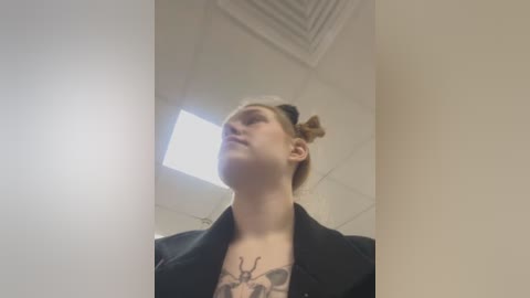 Media: Video of a young woman with fair skin, blonde hair in a bun, wearing a black blazer over a floral shirt with insect tattoos. She stands in a white-walled room with fluorescent lighting, looking upwards.