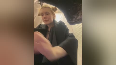 Media: A blurred video shows a young woman with blonde hair in two buns, wearing a black jacket, sitting on a bed with a black lace lingerie piece hanging on the wall.