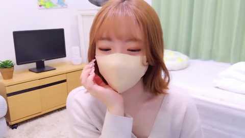 Media: Video of a young Asian woman with shoulder-length red hair and fair skin, wearing a beige face mask and white robe, holding a phone, in a light-colored bedroom with a green curtain, wooden desk, and a potted plant.