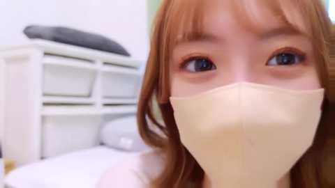 Media: Video of a young woman with fair skin, brown hair, and blue eyes wearing a surgical mask, standing in a blurred bedroom with white shelves and a bed.