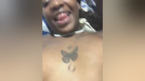 Media: Video of a topless Black woman with a butterfly tattoo on her chest, sticking out her tongue and wearing a choker. Background is blurred.