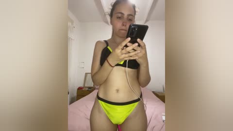 Media: A video of a young woman in bright yellow and black lingerie, standing in a modestly decorated bedroom, taking a selfie with a smartphone.