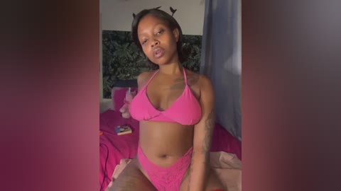 A video of a young Black woman with medium-brown skin, wearing a pink bikini, sitting on a bed with pink sheets. The room has dark green wall decor and a window with blue curtains.