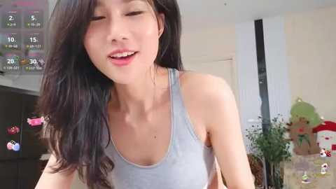 Media: Video of an Asian woman with long black hair, fair skin, and small breasts, wearing a light gray tank top, smiling, in a cozy, brightly lit room with colorful decorations.