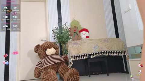 A video of a cozy room with a plush brown teddy bear, a green stuffed animal, a plant, and a quilted bedspread, against striped wallpaper and a door.