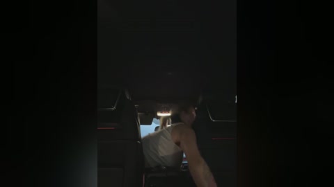 Media: Video of a dimly lit car interior, focusing on a man in a white tank top and dark pants, sitting with his head down, facing the rear. The background is dark and shadowy, making the subject stand out.