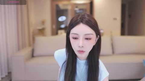 Media: Video of a young Asian woman with pale skin, long straight black hair, and a light blue turtleneck sweater, sitting indoors in a modern living room with beige sofas and wooden furniture.