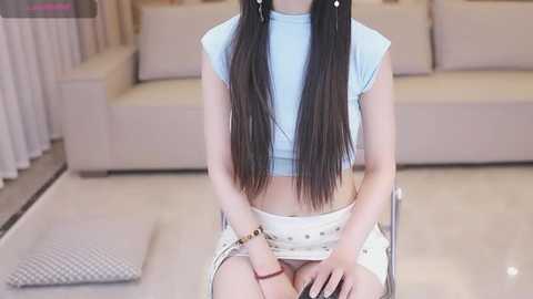 Media: Video of an Asian woman with long black hair, wearing a light blue crop top and white skirt, seated on a chair in a modern living room with beige sofa and white curtains.