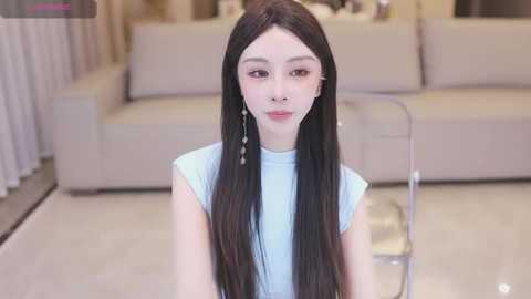 Media: Video of a young Asian woman with long, straight black hair, fair skin, and wearing a light blue sleeveless top, sitting indoors on a shiny white floor. Background features a beige sofa and abstract art.