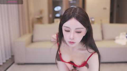 Media: A video of an Asian woman with long black hair, wearing a red bra, adjusting her straps in a modern living room with beige couches and neutral walls.