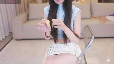 Media: A video of a young Asian woman with long black hair, sitting on a white couch, wearing a sleeveless light blue top and white shorts, playing with a yellow toy.